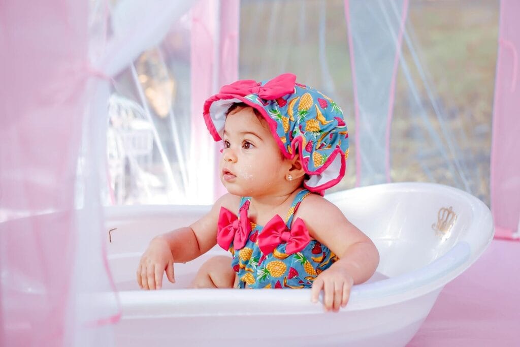 Cute baby sitting in a bathtub wearing a colorful dress and hat, surrounded by pink canopy.
