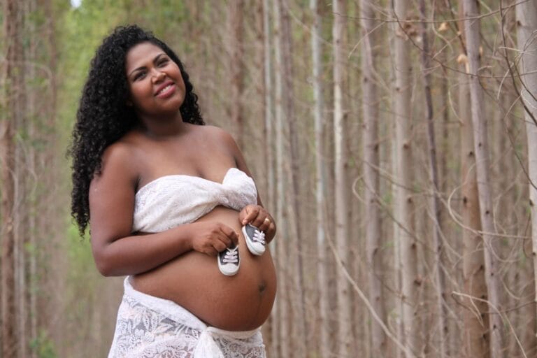 Smiling pregnant woman holding baby shoes in a serene forest setting.
